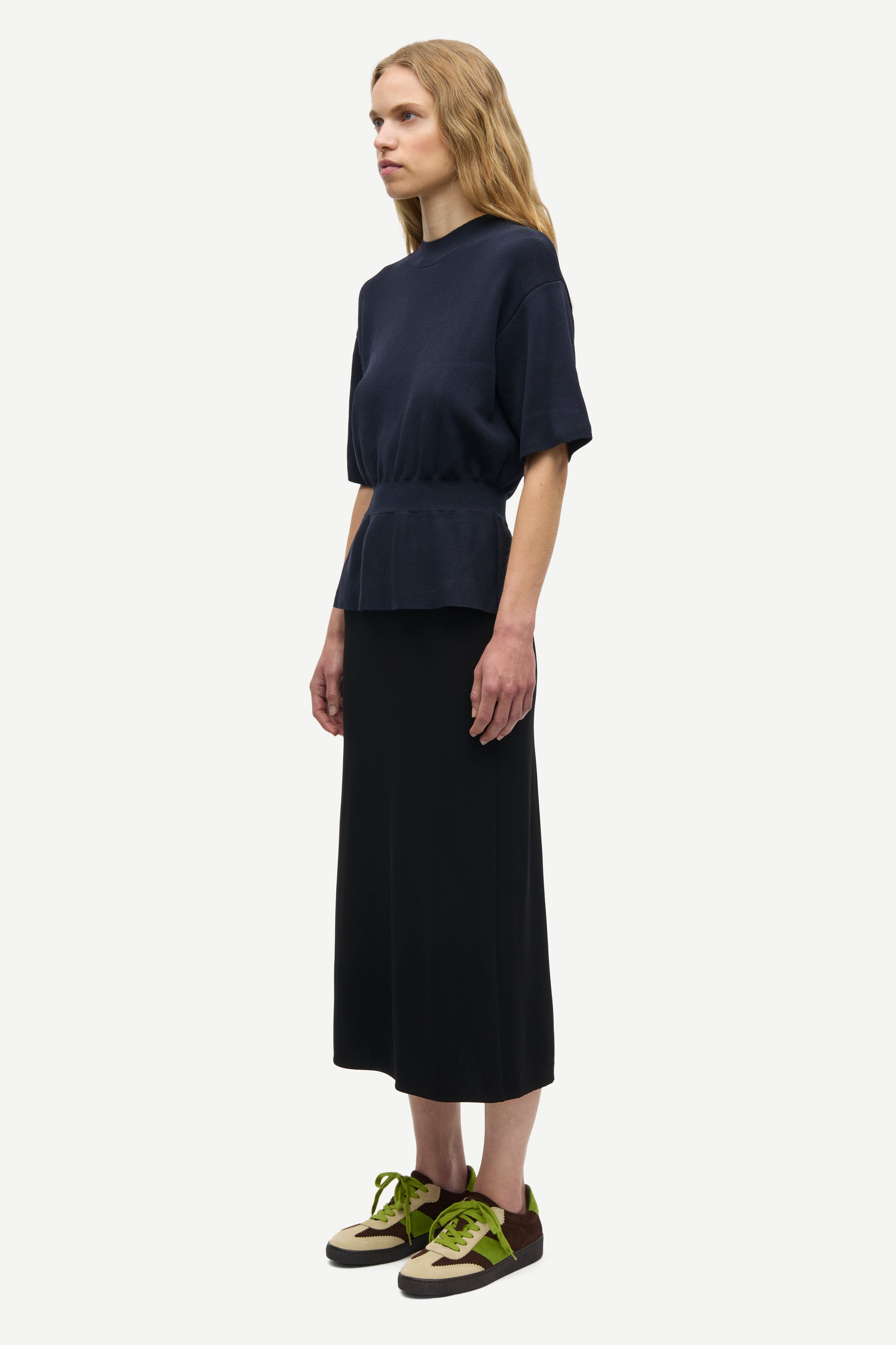 A model is shown from the side wearing a dark navy blue short-sleeve top with a peplum waist, a long black skirt, and olive green sneakers against a plain light gray background.