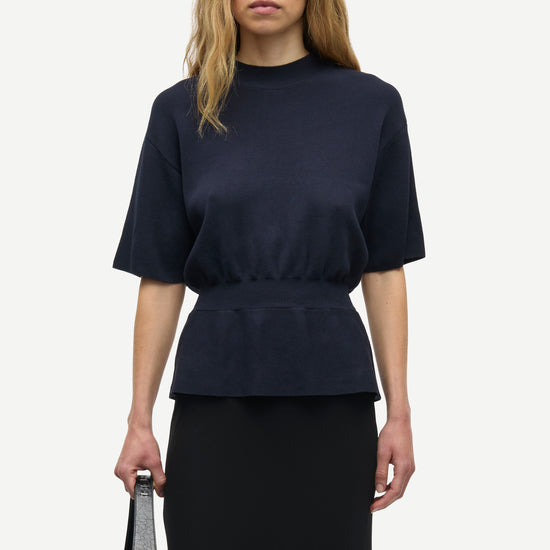 A woman modeling a dark navy blue short-sleeve top with a gathered waist, paired with a black skirt, holding a small gray handbag against a light gray background.