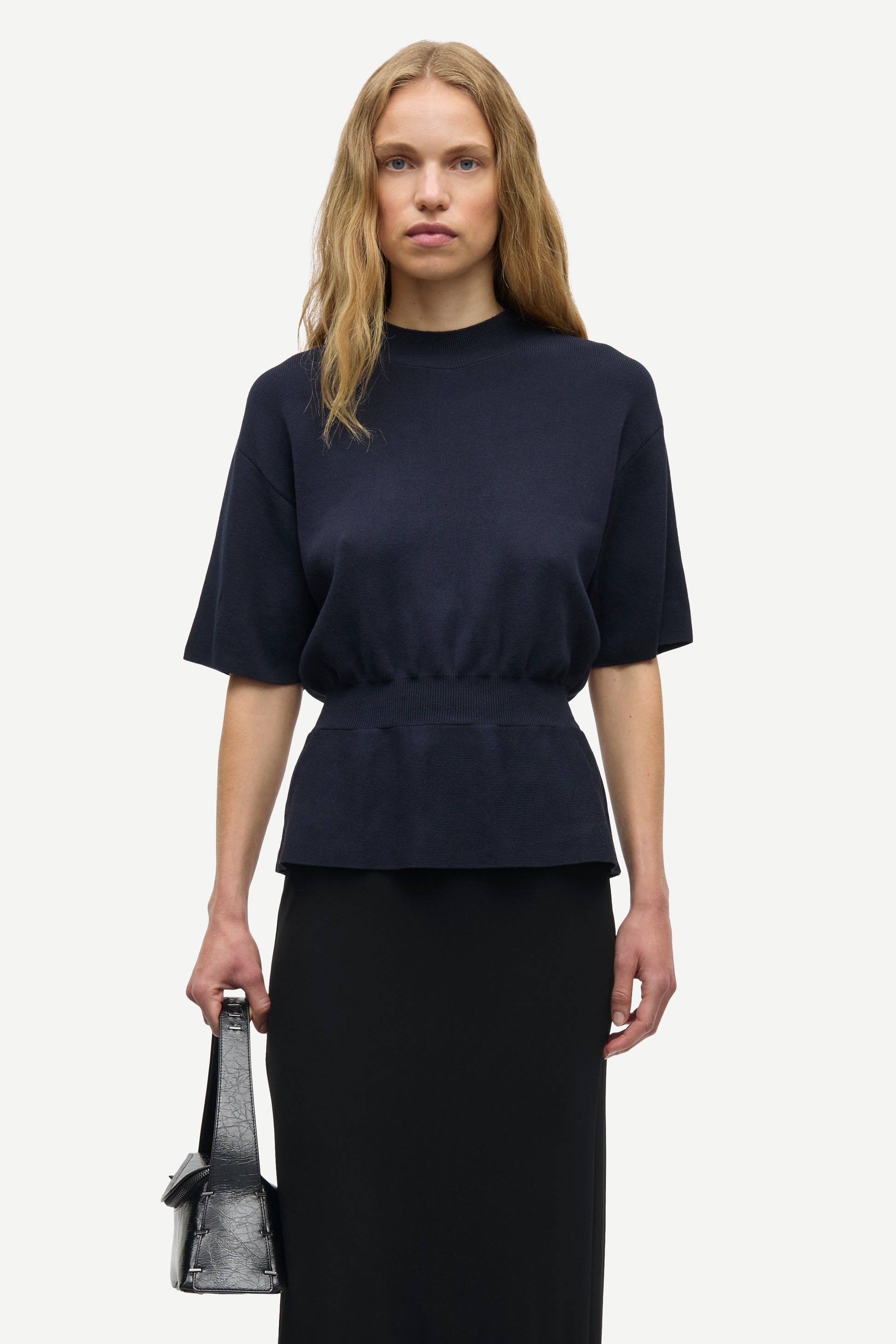 A woman modeling a dark navy blue short-sleeve top with a gathered waist, paired with a black skirt, holding a small gray handbag against a light gray background.