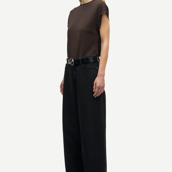 A model stands in profile wearing a dark brown short-sleeve top, black wide-leg trousers, and a black belt with a silver buckle, against a plain light gray studio background.