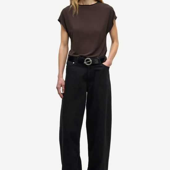 A woman modeling a dark brown short-sleeve crewneck t-shirt and black wide-leg trousers with a black belt, standing against a light gray studio background.