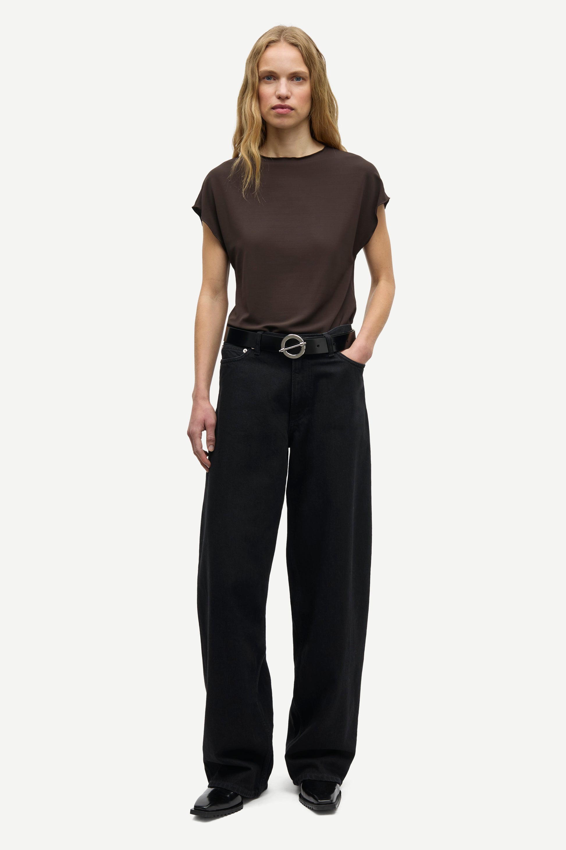 A woman modeling a dark brown short-sleeve crewneck t-shirt and black wide-leg trousers with a black belt, standing against a light gray studio background.