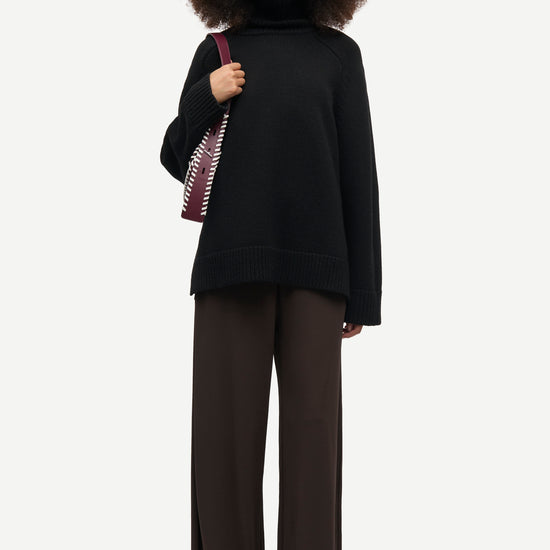 A model is wearing a black oversized mock-neck sweater, dark brown wide-leg trousers, and black shoes, while carrying a patterned shoulder bag against a light gray background.