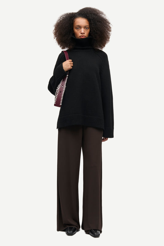 A model is wearing a black oversized mock-neck sweater, dark brown wide-leg trousers, and black shoes, while carrying a patterned shoulder bag against a light gray background.