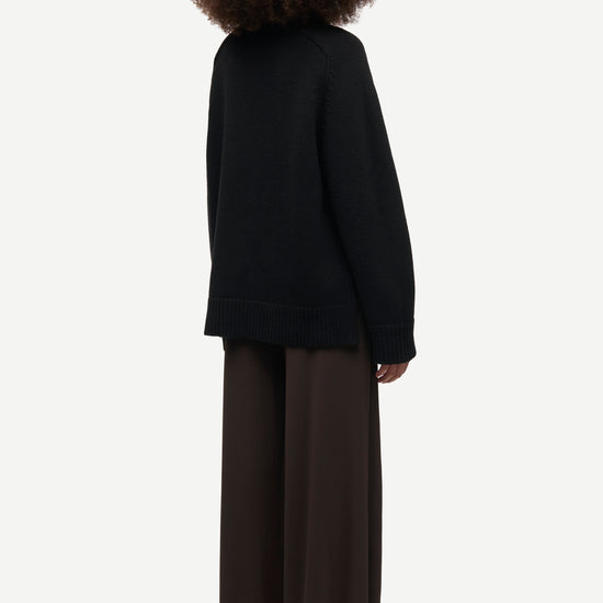 A person shown from the back wearing a black knit sweater and dark brown wide-leg trousers against a plain light gray studio background.