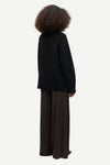 A person shown from the back wearing a black knit sweater and dark brown wide-leg trousers against a plain light gray studio background.