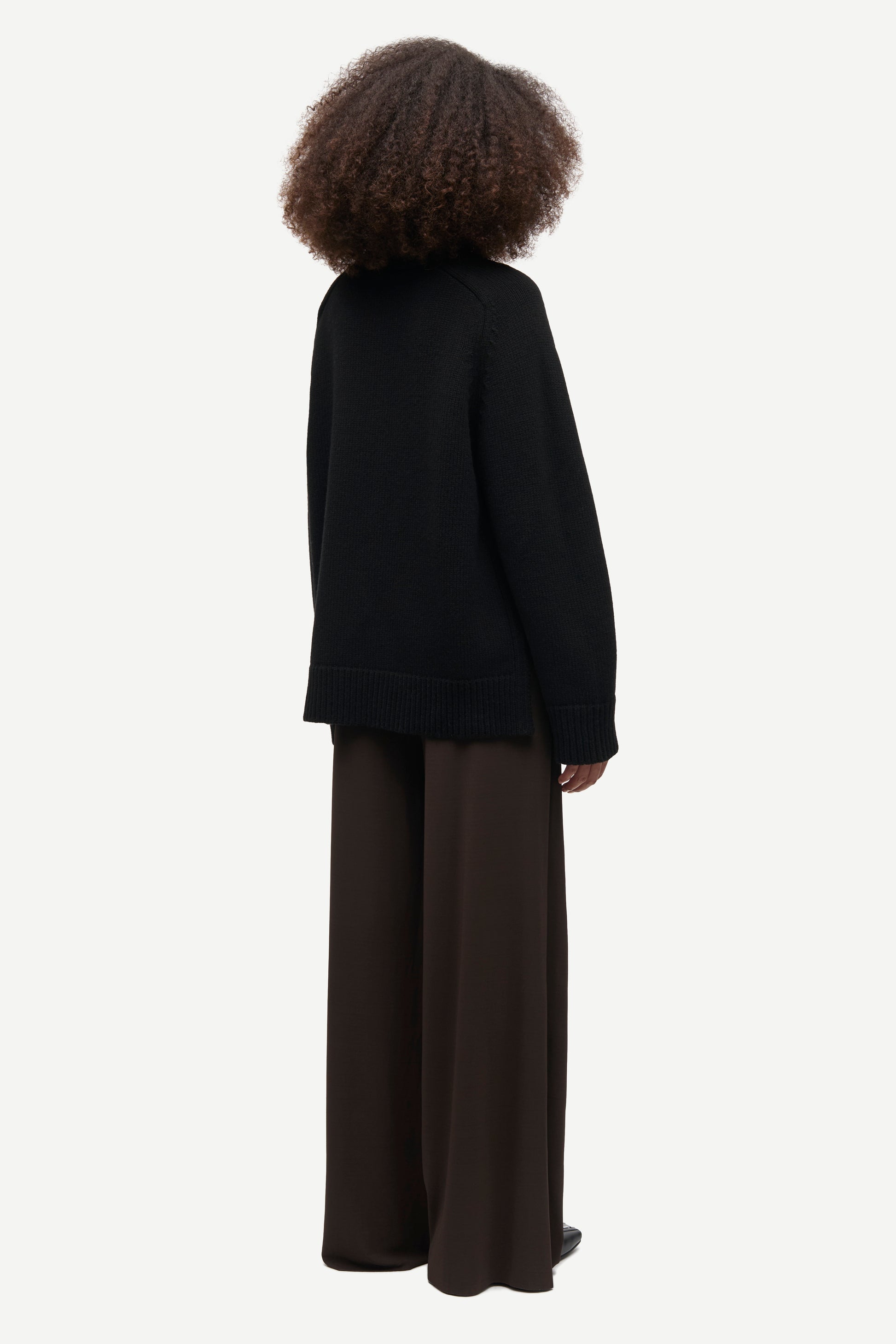 A person shown from the back wearing a black knit sweater and dark brown wide-leg trousers against a plain light gray studio background.