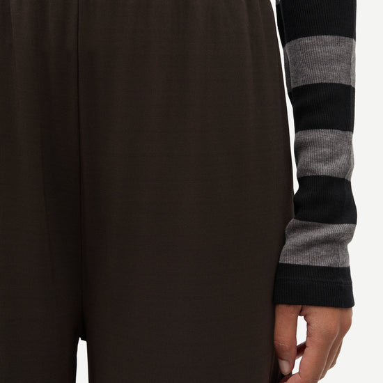 Close-up of a person wearing dark brown wide-leg trousers and a gray and black striped long-sleeve top, with the hand holding the hem of the pants visible against a plain light background.