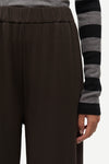 Close-up of a person wearing dark brown wide-leg trousers and a gray and black striped long-sleeve top, with the hand holding the hem of the pants visible against a plain light background.