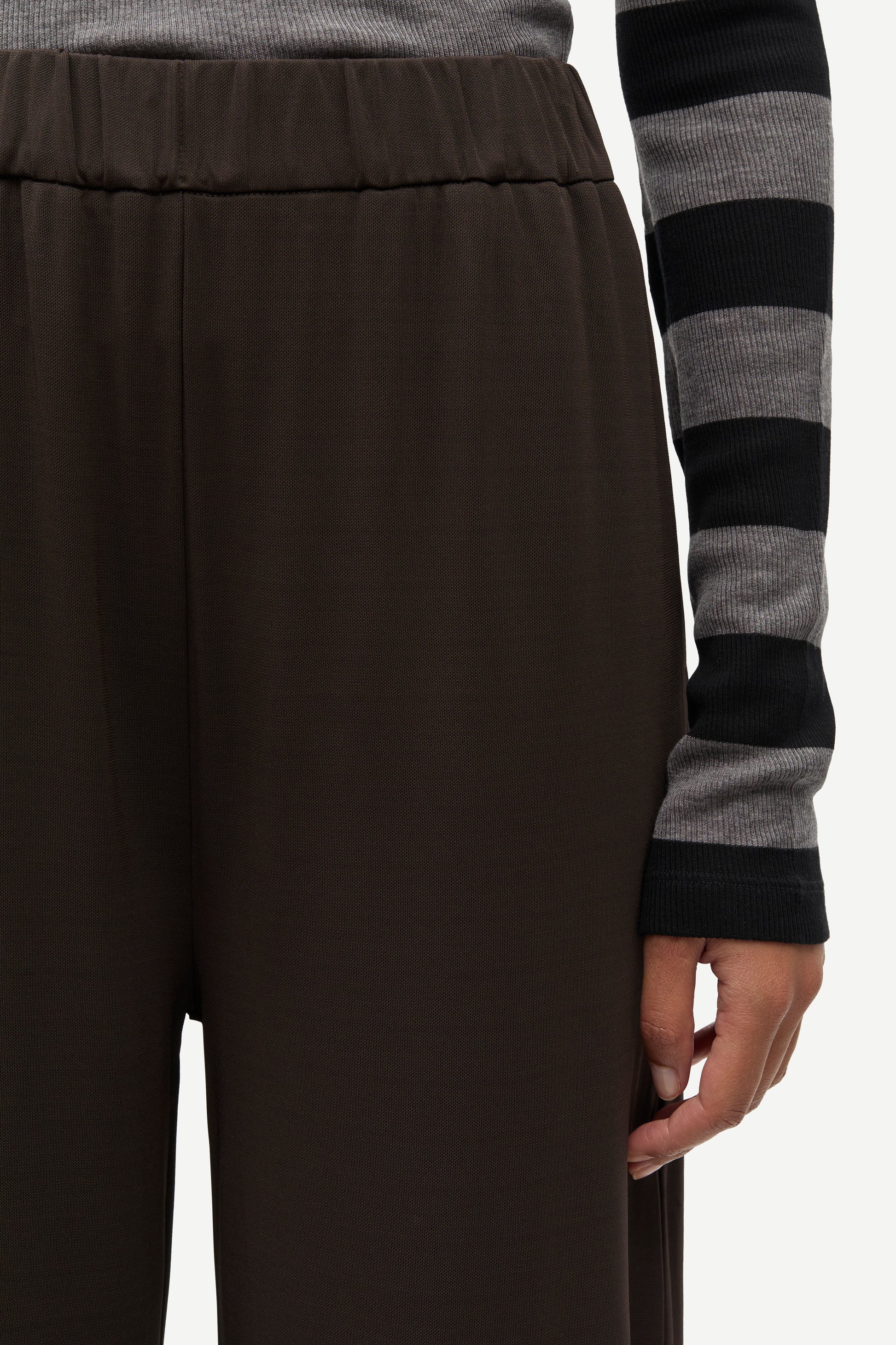 Close-up of a person wearing dark brown wide-leg trousers and a gray and black striped long-sleeve top, with the hand holding the hem of the pants visible against a plain light background.