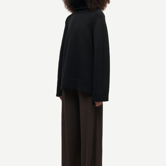 A model with voluminous dark curly hair is shown in profile wearing a black oversized turtleneck sweater and dark brown wide-leg trousers against a light gray studio background.