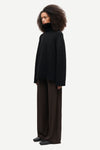 A model with voluminous dark curly hair is shown in profile wearing a black oversized turtleneck sweater and dark brown wide-leg trousers against a light gray studio background.