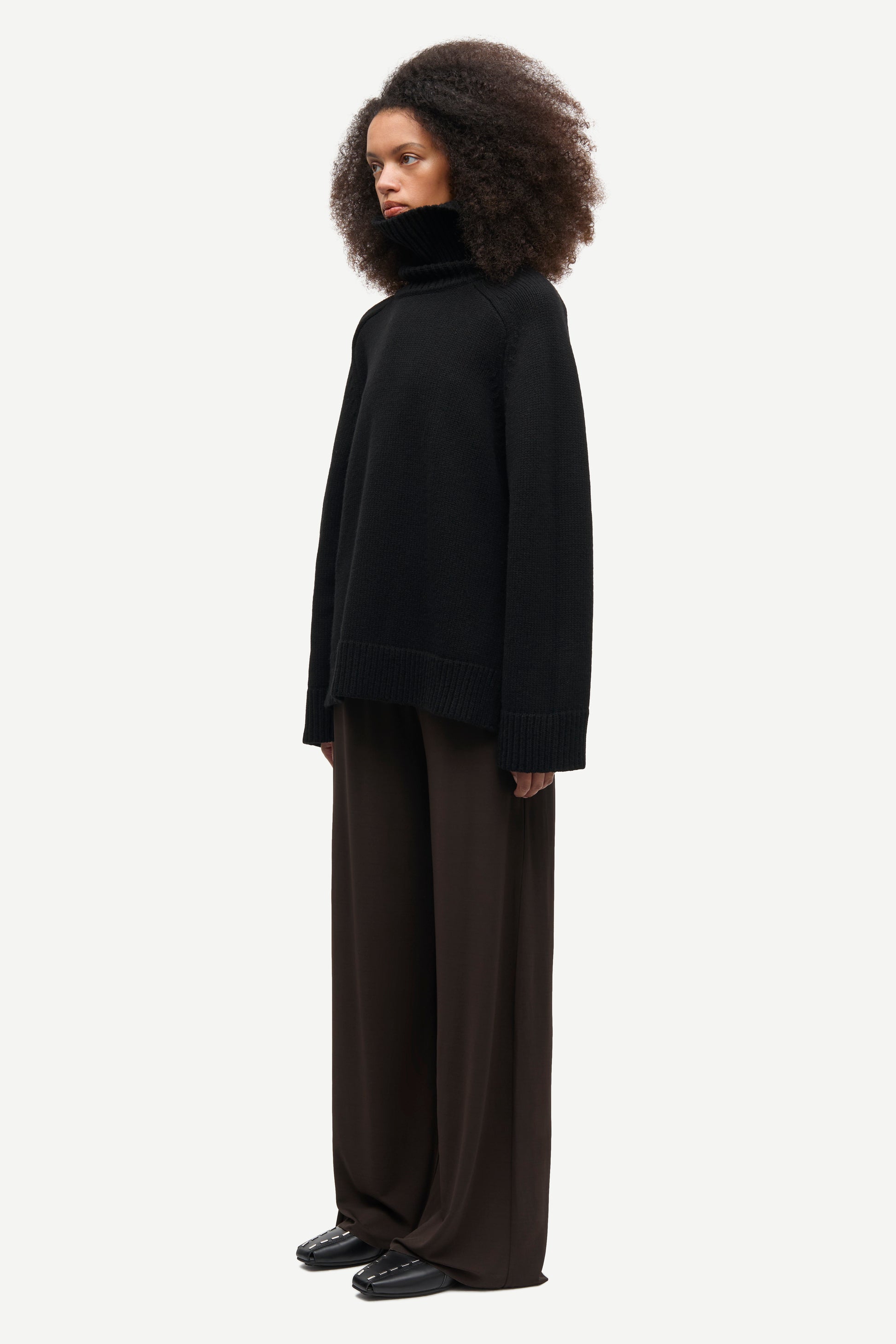 A model with voluminous dark curly hair is shown in profile wearing a black oversized turtleneck sweater and dark brown wide-leg trousers against a light gray studio background.