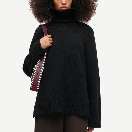 A model with voluminous dark curly hair wearing a black oversized turtleneck sweater and dark brown trousers, holding a burgundy shoulder bag with a white patterned strap, against a light gray studio background.