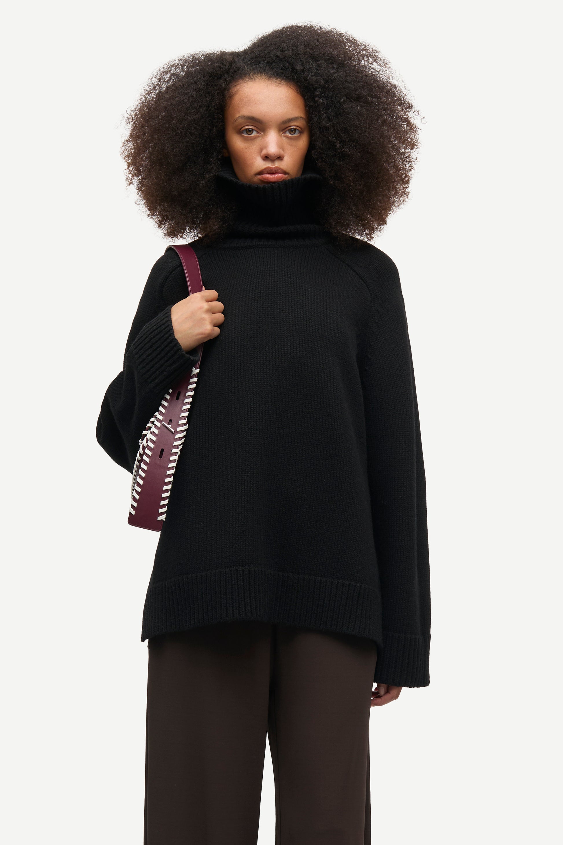 A model with voluminous dark curly hair wearing a black oversized turtleneck sweater and dark brown trousers, holding a burgundy shoulder bag with a white patterned strap, against a light gray studio background.