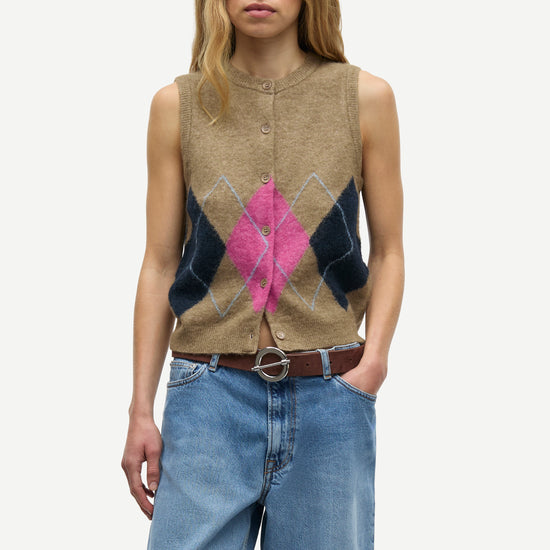A woman modeling a sleeveless button-front knit vest with an argyle pattern in beige, dark navy, and bright pink, paired with blue jeans and a brown belt against a light gray studio background.