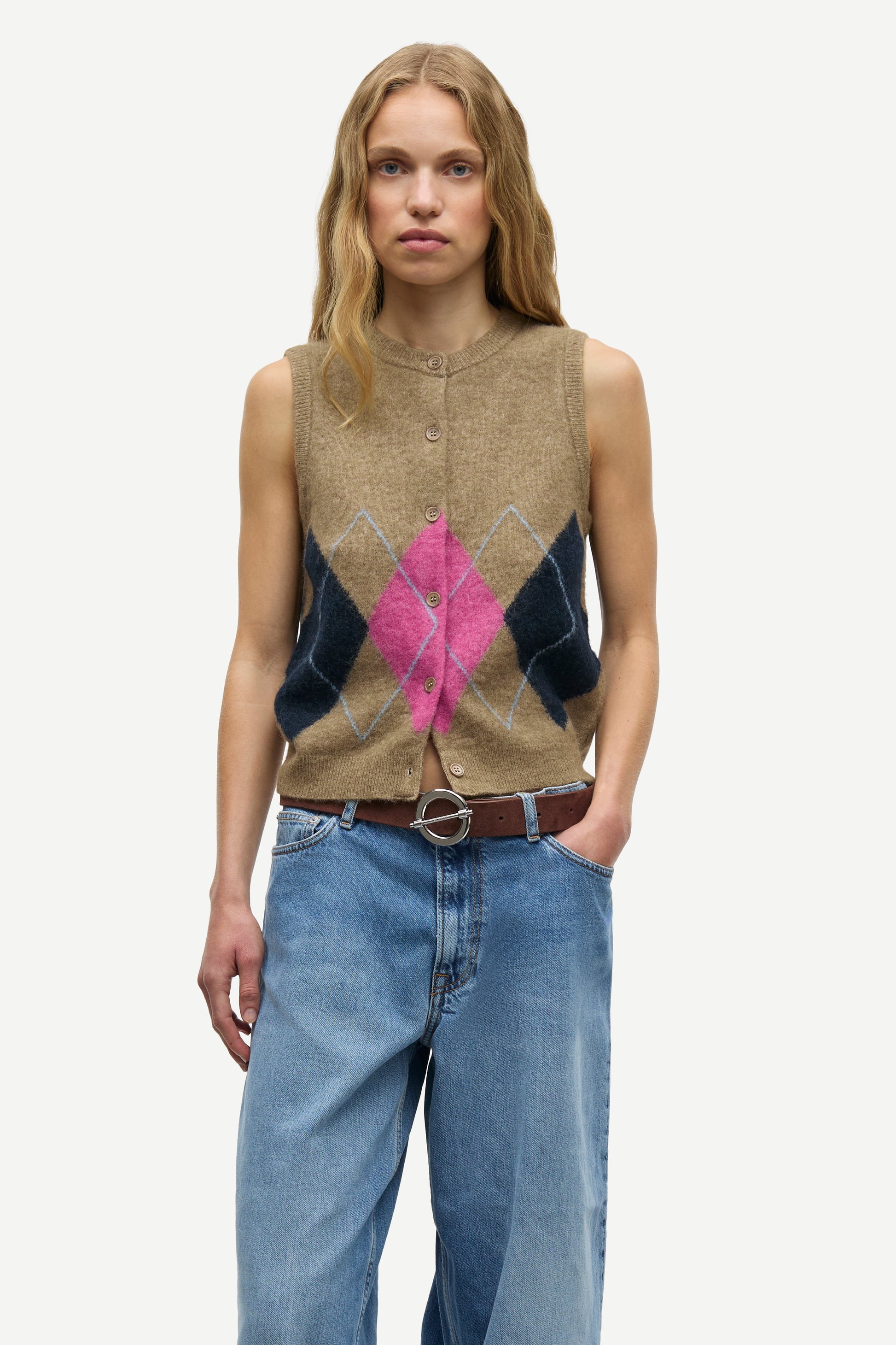 A woman modeling a sleeveless button-front knit vest with an argyle pattern in beige, dark navy, and bright pink, paired with blue jeans and a brown belt against a light gray studio background.