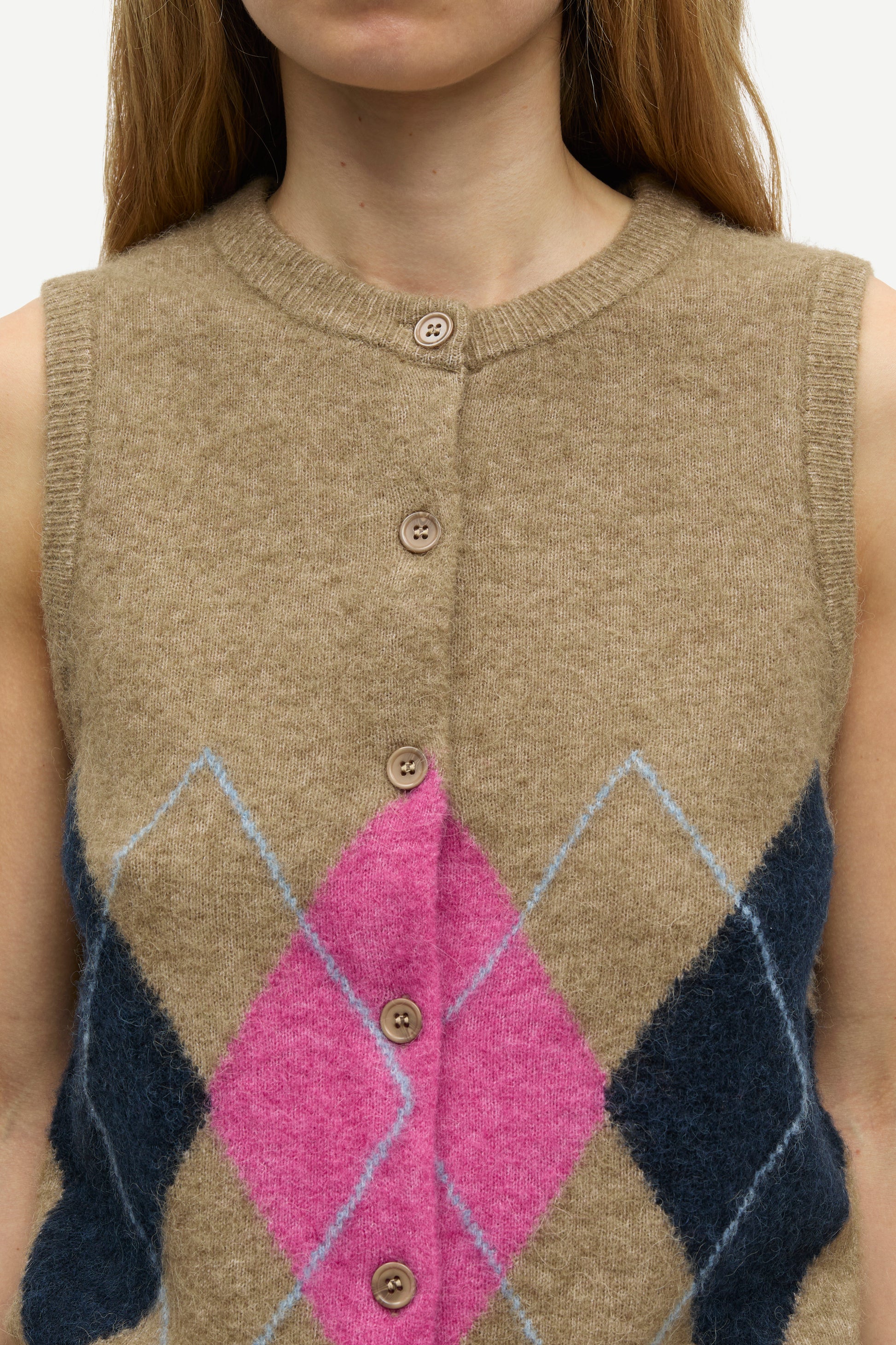 A close-up of a person wearing a sleeveless knit button-front vest in beige with a large pink diamond and navy blue argyle-style geometric pattern on the lower front.