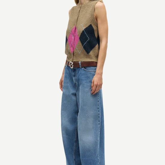 A woman stands in profile wearing a sleeveless beige knit vest with a pink and dark blue diamond pattern, wide-leg blue jeans, a brown belt, and black shoes against a light gray studio background.