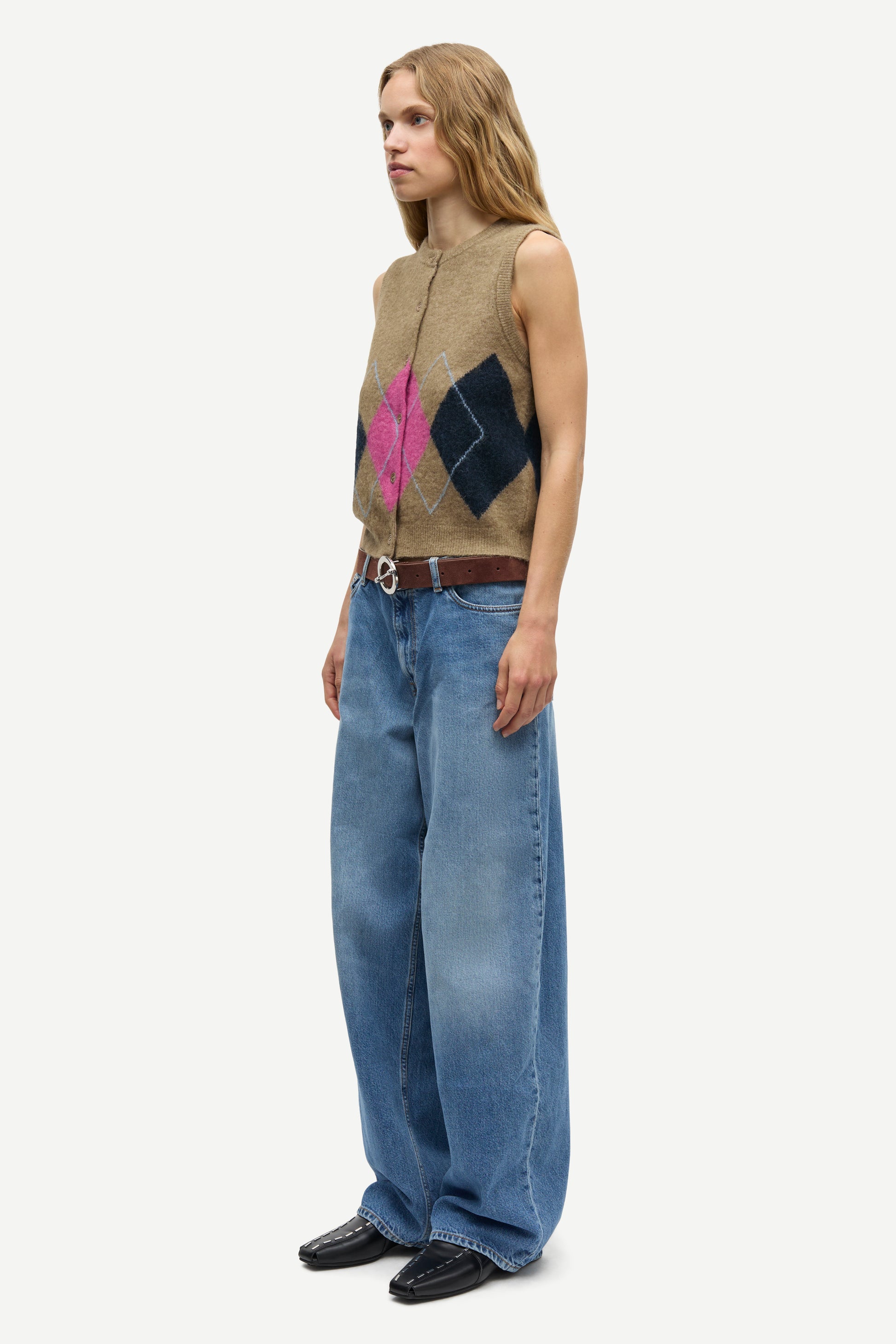 A woman stands in profile wearing a sleeveless beige knit vest with a pink and dark blue diamond pattern, wide-leg blue jeans, a brown belt, and black shoes against a light gray studio background.