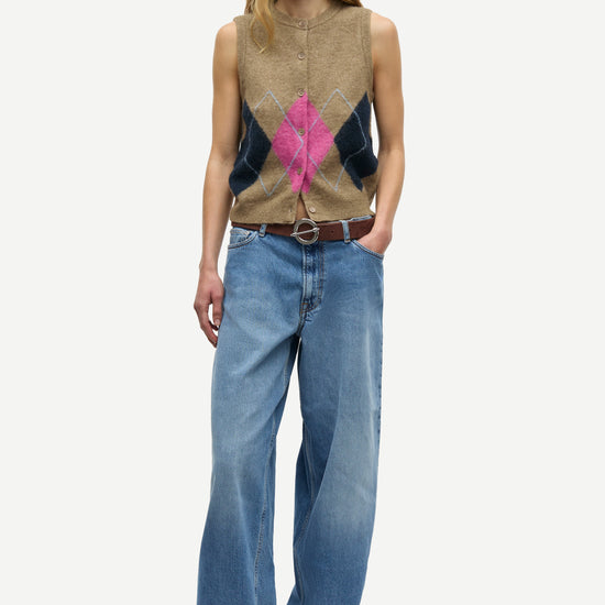 A model stands against a light gray studio background wearing a sleeveless knit sweater vest with a tan base, dark navy side panels, and a bright pink geometric pattern at the center, paired with wide-leg blue jeans and black shoes.