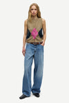 A model stands against a light gray studio background wearing a sleeveless knit sweater vest with a tan base, dark navy side panels, and a bright pink geometric pattern at the center, paired with wide-leg blue jeans and black shoes.
