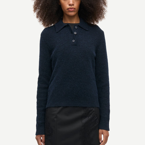 A model with curly dark hair wearing a dark navy blue long-sleeve knit polo sweater and a black mini skirt, standing against a light gray studio background.