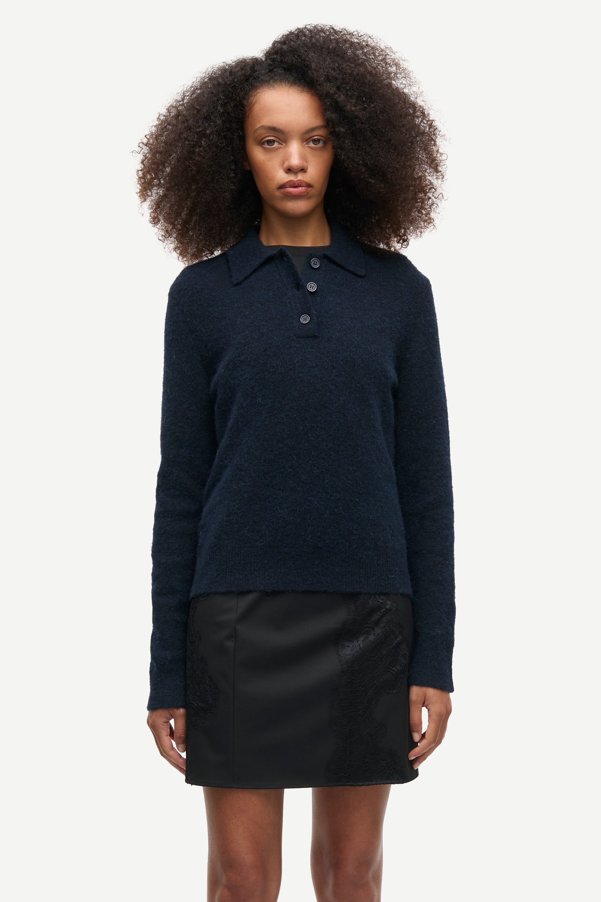 A model with curly dark hair wearing a dark navy blue long-sleeve knit polo sweater and a black mini skirt, standing against a light gray studio background.