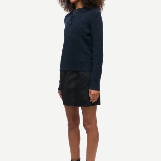 A model wearing a dark navy blue knit polo sweater, a black mini skirt, and black leather ankle boots, shown in profile against a light gray studio background.
