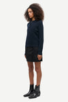 A model wearing a dark navy blue knit polo sweater, a black mini skirt, and black leather ankle boots, shown in profile against a light gray studio background.