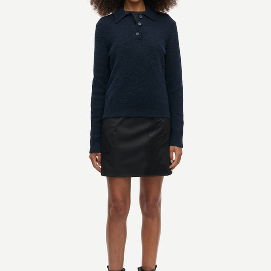 A model stands against a light gray studio background wearing a dark navy long-sleeve knit polo sweater, a black mini skirt, and black ankle boots.
