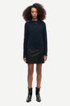 A model stands against a light gray studio background wearing a dark navy long-sleeve knit polo sweater, a black mini skirt, and black ankle boots.