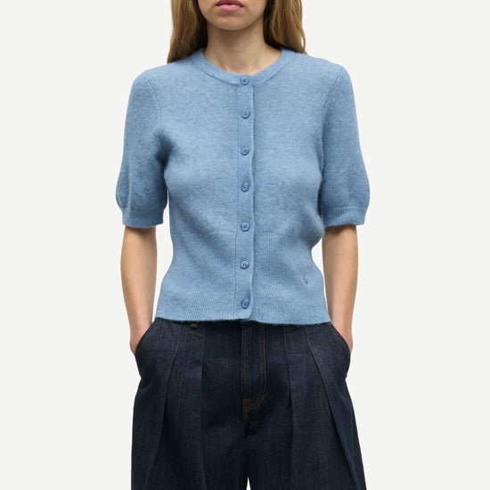 A woman modeling a light blue short-sleeve button-front cardigan with a crew neck, paired with dark wide-leg trousers, against a plain light gray studio background.