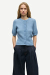 A woman modeling a light blue short-sleeve button-front cardigan with a crew neck, paired with dark wide-leg trousers, against a plain light gray studio background.