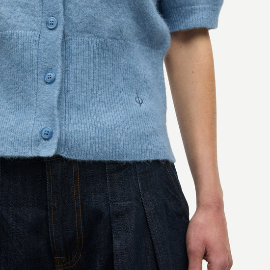 Close-up of a person wearing a light blue short-sleeve knit cardigan with blue buttons and dark pleated trousers, against a plain light background.