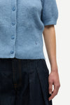 Close-up of a person wearing a light blue short-sleeve knit cardigan with blue buttons and dark pleated trousers, against a plain light background.