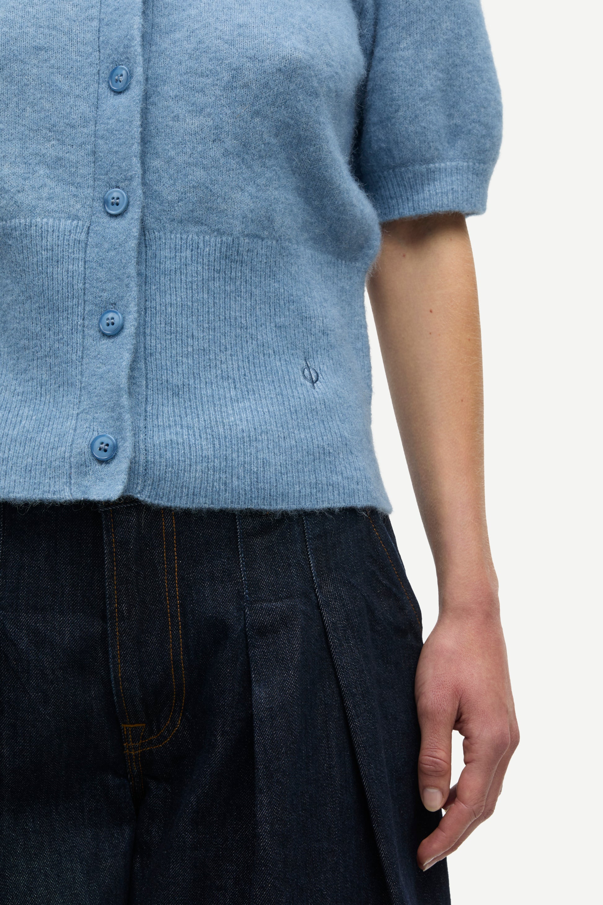 Close-up of a person wearing a light blue short-sleeve knit cardigan with blue buttons and dark pleated trousers, against a plain light background.