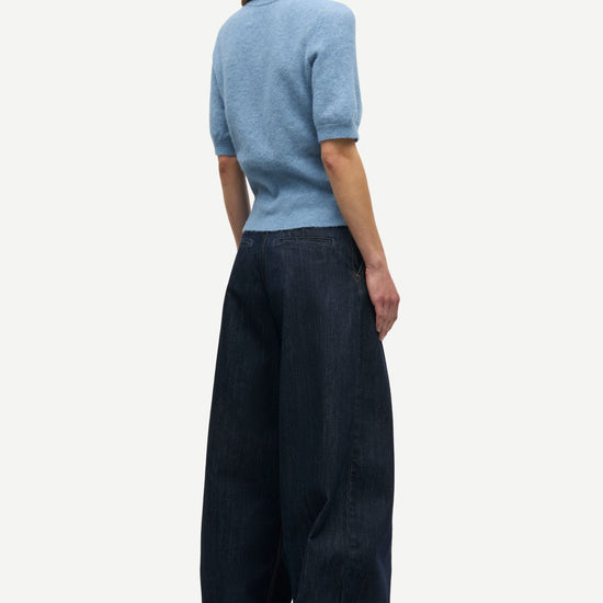 A woman shown from the back wearing a light blue short-sleeve knit top and dark wide-leg trousers, photographed against a plain light gray studio background.
