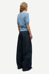 A woman shown from the back wearing a light blue short-sleeve knit top and dark wide-leg trousers, photographed against a plain light gray studio background.
