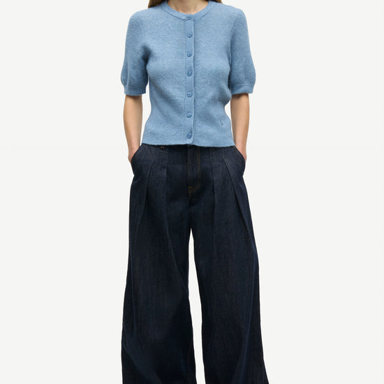 A woman modeling a light blue short-sleeve button-front cardigan with dark wide-leg trousers, standing against a plain light gray studio background.