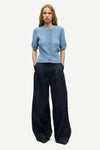 A woman modeling a light blue short-sleeve button-front cardigan with dark wide-leg trousers, standing against a plain light gray studio background.