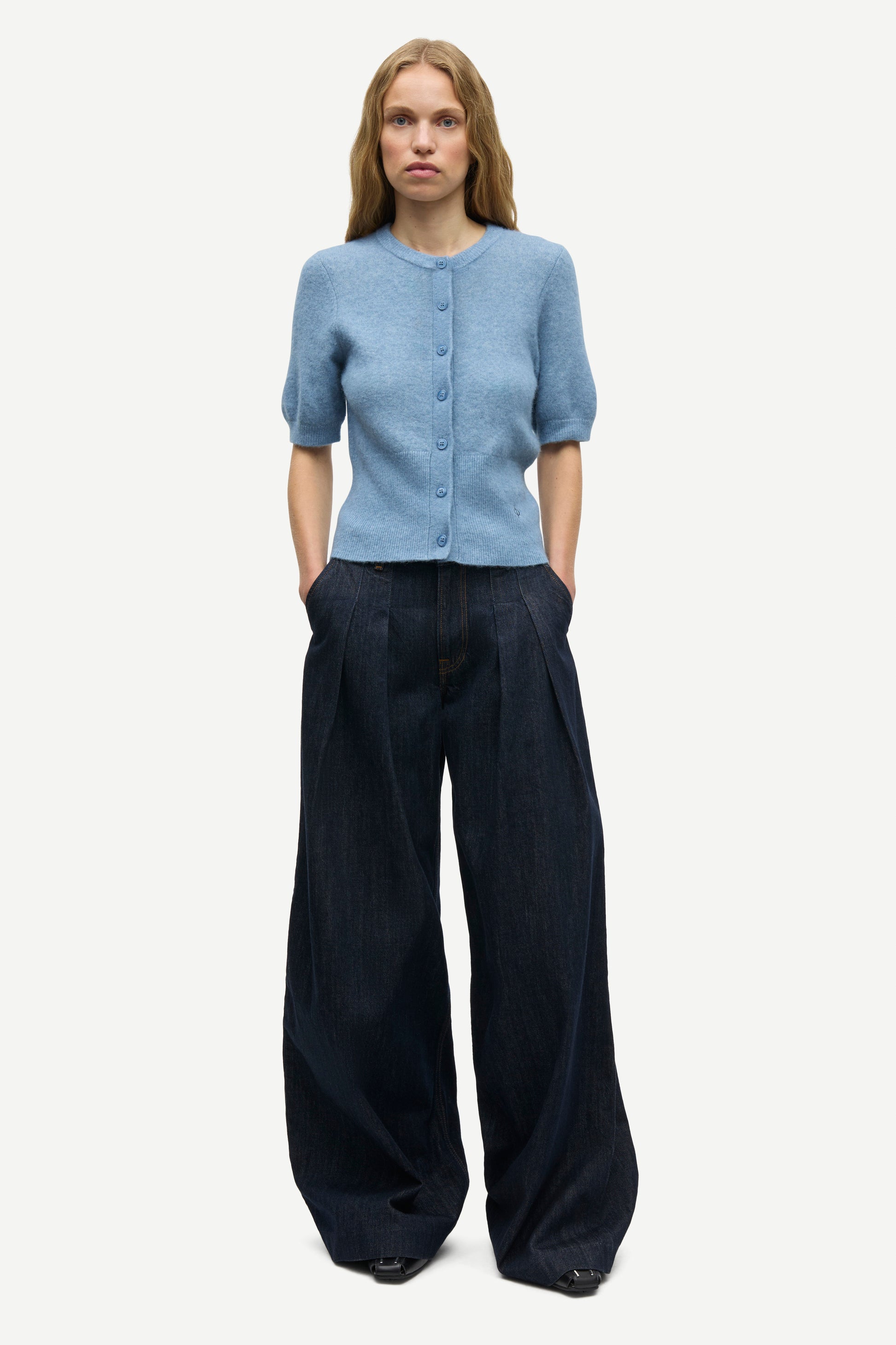 A woman modeling a light blue short-sleeve button-front cardigan with dark wide-leg trousers, standing against a plain light gray studio background.