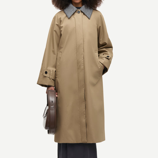 A model with curly dark hair stands against a light gray studio background wearing a long beige trench coat with a contrasting gray collar, dark wide-leg skirt, and holding a brown leather circular case.
