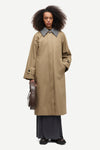 A model with curly dark hair stands against a light gray studio background wearing a long beige trench coat with a contrasting gray collar, dark wide-leg skirt, and holding a brown leather circular case.