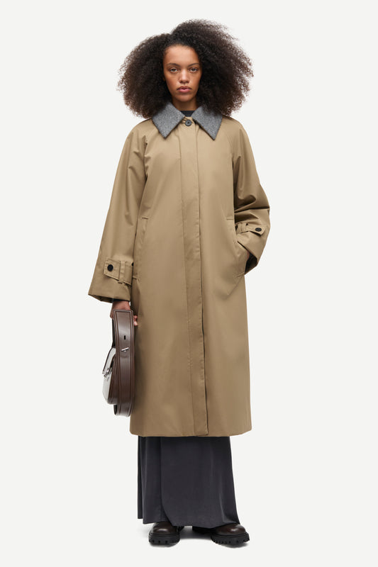 A model with curly dark hair stands against a light gray studio background wearing a long beige trench coat with a contrasting gray collar, dark wide-leg skirt, and holding a brown leather circular case.
