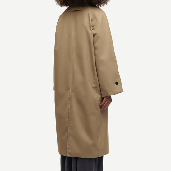 A model shown from the back wearing a long beige trench coat over dark gray wide-leg trousers and black shoes, set against a plain light gray background.