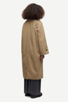 A model shown from the back wearing a long beige trench coat over dark gray wide-leg trousers and black shoes, set against a plain light gray background.