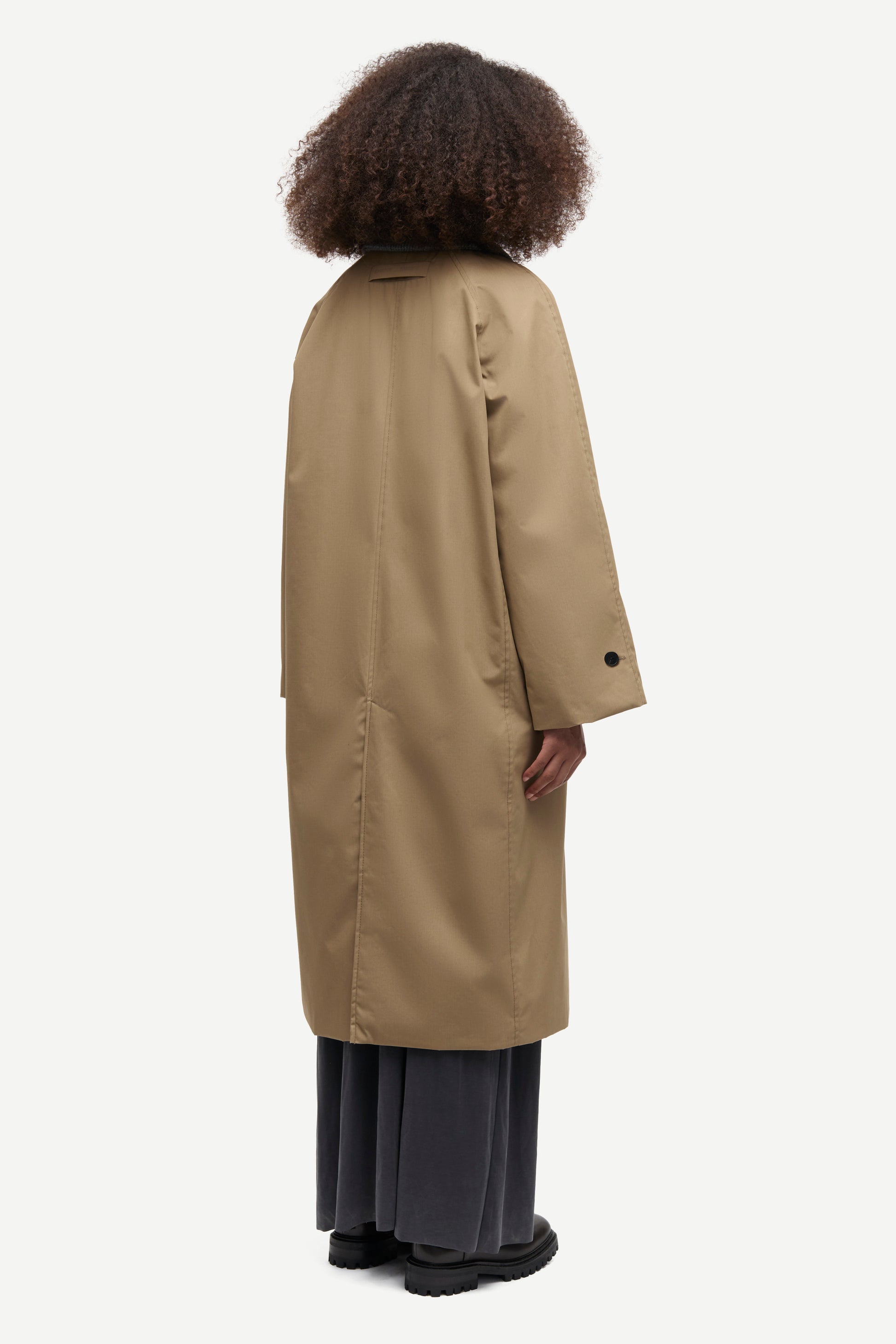 A model shown from the back wearing a long beige trench coat over dark gray wide-leg trousers and black shoes, set against a plain light gray background.