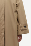 Close-up of a beige trench coat sleeve with a buttoned cuff detail, showing part of a person's hand at the hem against a light gray background.