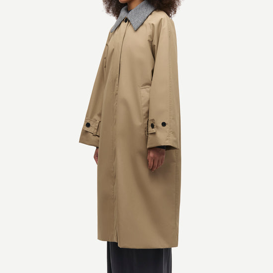 A model wearing a mid-length beige trench coat with a contrasting gray collar, paired with dark wide-leg trousers and black shoes, shown from a side profile against a light gray background.